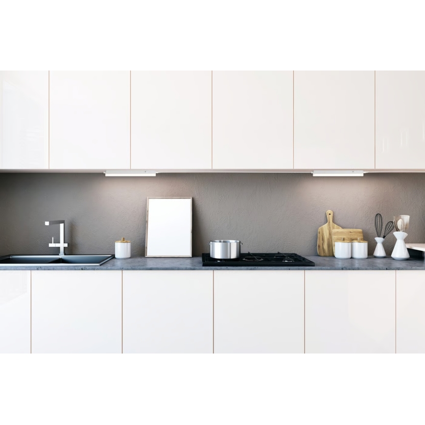 Kuchynská linka s bielymi skrinkami a sivou pracovnou doskou. Na doske je umiestnený hrniec, nádoba, rám obrazu a kuchynské náčinie. Pod skrinkami je osvetlenie.