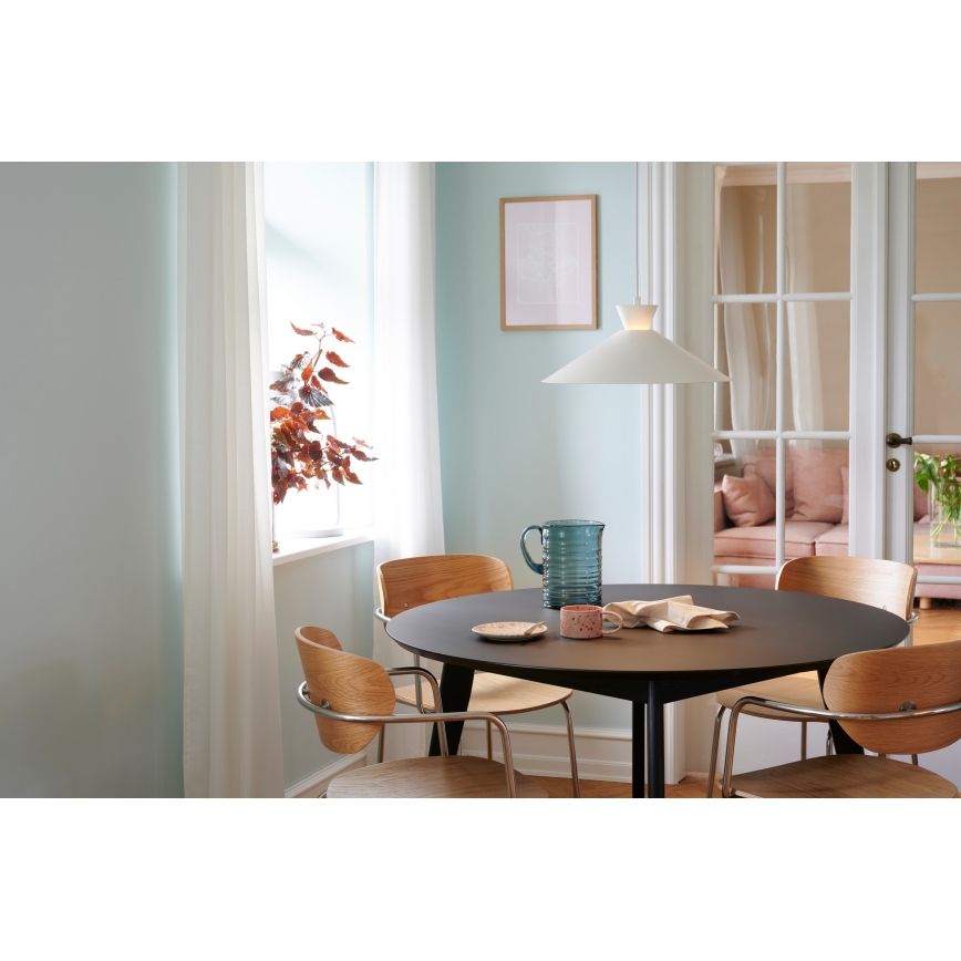 Okrúhly stôl s drevenými stoličkami je prestretý džbánom a hrnčekom. Nad stolom visí biela lampa. Prostredie tvorí svetlá izba s oknom a dverami do ďalšej miestnosti.