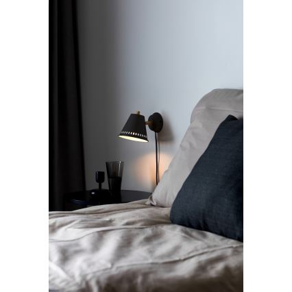 Čierna nástenná lampa s kužeľovitým tienidlom s otvormi svieti v spálni pri posteli s posteľnou bielizňou a vankúšmi. Lampa je pripevnená na stene vedľa nočného stolíka.