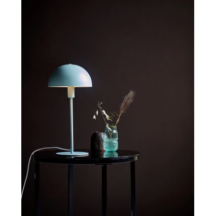 Stolová lampa s bledomodrým tienidlom v tvare huby stojí na čiernom stolíku. Na stolíku je aj váza so suchou kvetinou a malá dekorácia. Pozadie je tmavohnedé.