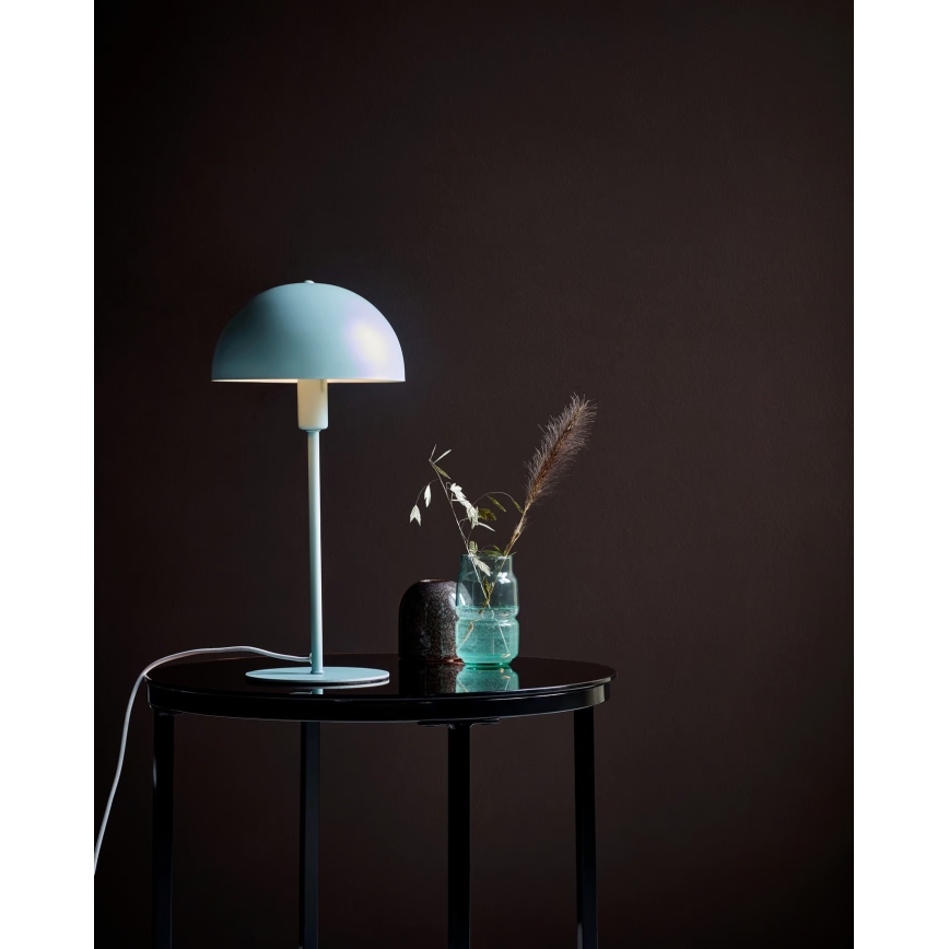 Stolová lampa s bledomodrým tienidlom v tvare huby stojí na čiernom stolíku. Na stolíku je aj váza so suchou kvetinou a malá dekorácia. Pozadie je tmavohnedé.
