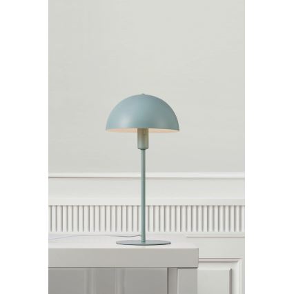 Stolová lampa svetlomodrej farby s polguľovitým tienidlom stojí na bielom stole pred bielou stenou s lištami.