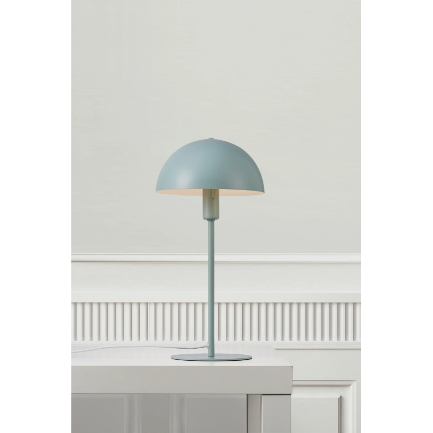 Stolová lampa svetlomodrej farby s polguľovitým tienidlom stojí na bielom stole pred bielou stenou s lištami.
