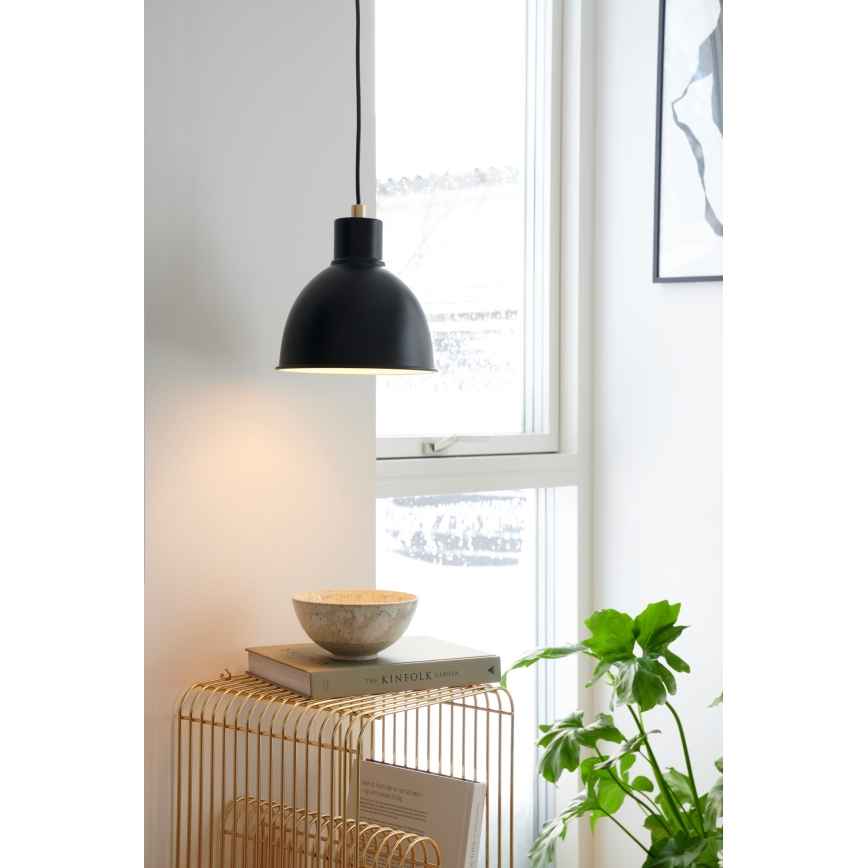Čierna závesná lampa svieti v interiéri. Na zlatom stolíku pod lampou leží miska a kniha s názvom „The Kinfolk Garden“. V pozadí je okno a zelená rastlina.