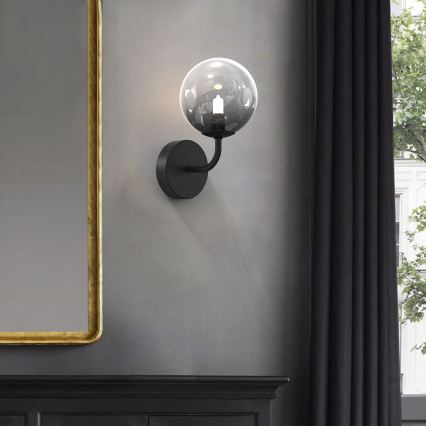 Čierna nástenná lampa s guľovou sklenenou tienidlom svieti. Lampa je pripevnená na sivej stene vedľa zrkadla a tmavej záclony.