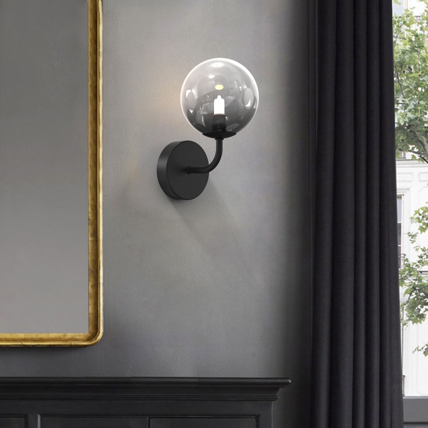 Čierna nástenná lampa s guľovou sklenenou tienidlom svieti. Lampa je pripevnená na sivej stene vedľa zrkadla a tmavej záclony.