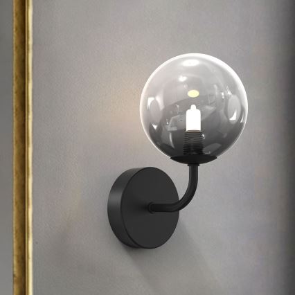 Nástená lampa s čiernym kovom a sklenenou guľou svieti na sivej stene. Svetlo pochádza z vnútra sklenenej gule. V pozadí je časť zlatej steny.