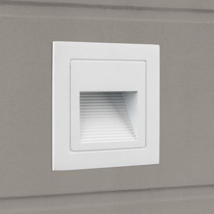 Biela nástenná LED lampa je zabudovaná do šedej steny. Lampa má štvorcový tvar s úzkym otvorom s vertikálnymi drážkami, ktorý vyžaruje svetlo.