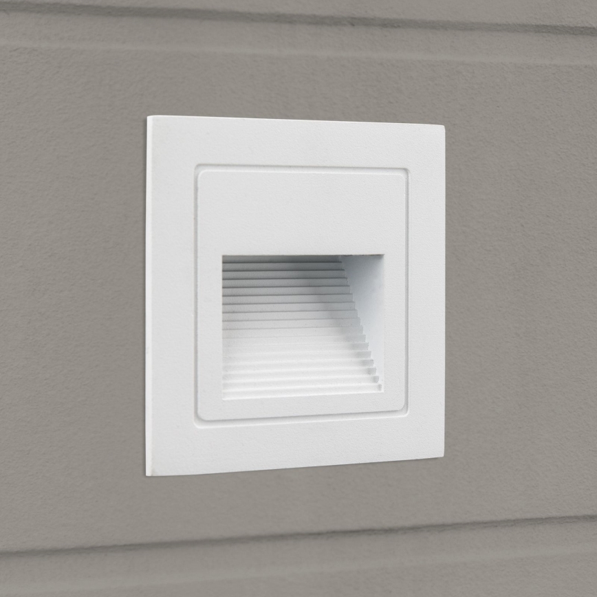 Biela nástenná LED lampa je zabudovaná do šedej steny. Lampa má štvorcový tvar s úzkym otvorom s vertikálnymi drážkami, ktorý vyžaruje svetlo.