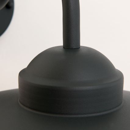 Čierna nástenná lampa je pripevnená na stenu. Lampa má zaoblený tvar s kupolovitou hornou časťou a zahnutým ramenom. Pozadie je svetlé.