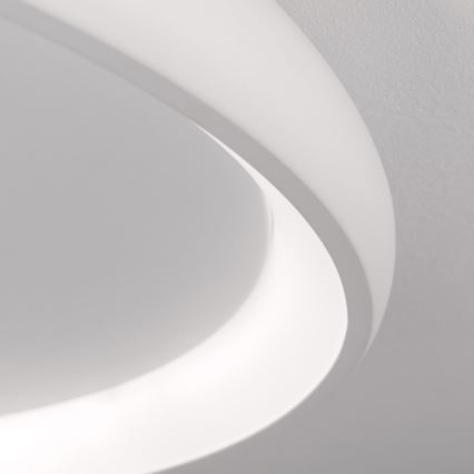 Biela stropná lampa svieti. Jej oválny tvar má vnútorný svetelný kruh. Lampa je pripevnená k svetlému stropu.