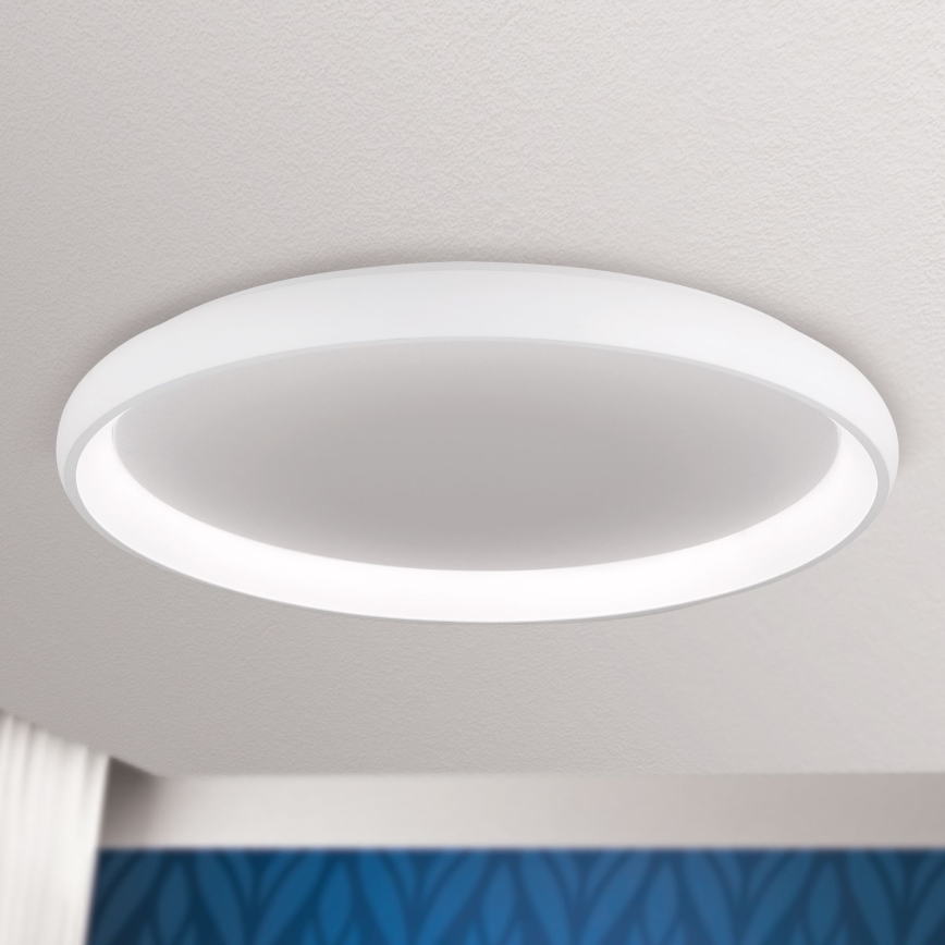 Biela kruhová stropná lampa svieti. Je pripevnená k stropu svetlej farby. Pod lampou je vidieť časť steny s tmavomodrým vzorom.
