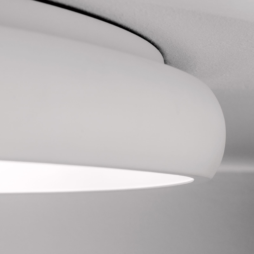 Biela stropná lampa je pripevnená k stropu. Svetlo svieti z vnútornej časti lampy. Lampa je zaobleného tvaru.