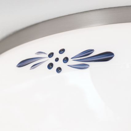 Detail biely porcelánový povrch s tmavomodrým vzorom listov a bobuliek. Vzor je umiestnený pod kovovými lemom.
