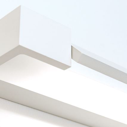 Biela nástenná lampa svieti. Je pripevnená k stene. Lampa má jednoduchý, geometrický tvar.