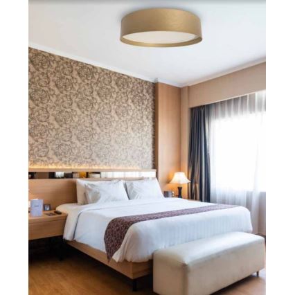 Stropné svietidlo svieti v spálni. Svietidlo je okrúhle, zlaté s bielym vnútorným difúzorom. Spálňa obsahuje manželskú posteľ, nočný stolík a svetlé drevené podlahy.