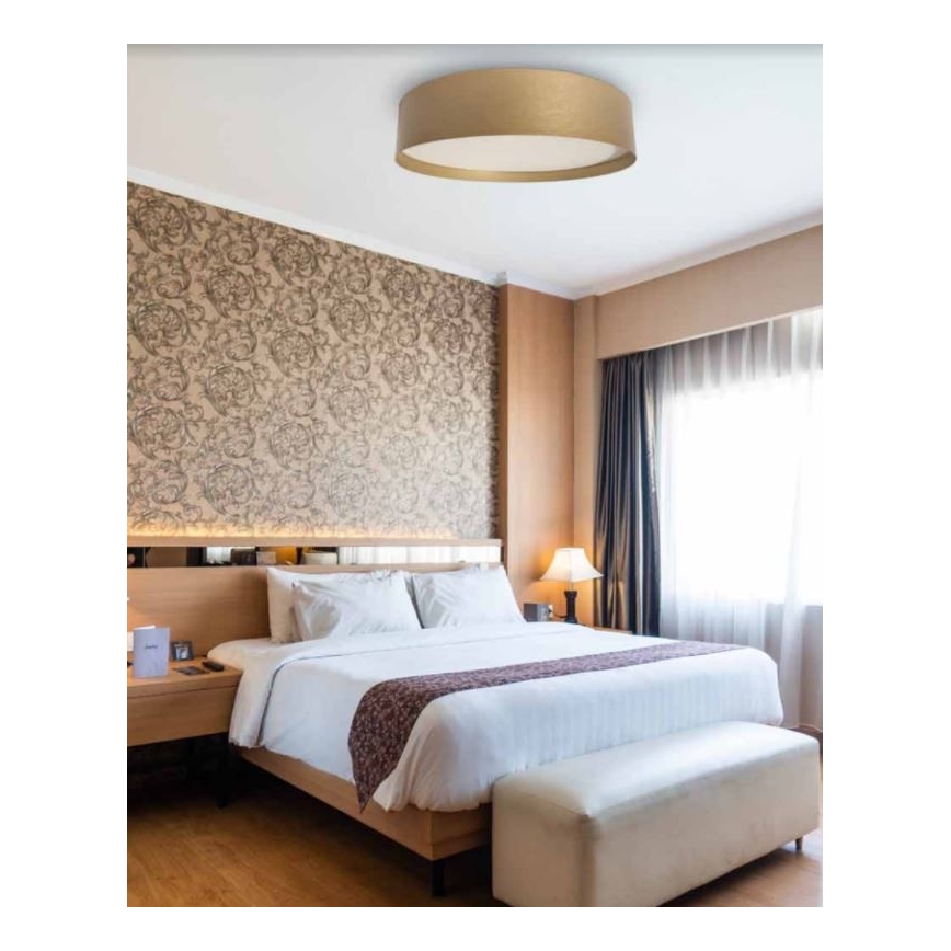 Stropné svietidlo svieti v spálni. Svietidlo je okrúhle, zlaté s bielym vnútorným difúzorom. Spálňa obsahuje manželskú posteľ, nočný stolík a svetlé drevené podlahy.