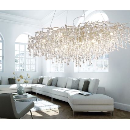 Veľký lustr s mnohými krištáľovými príveskami v tvare kvapiek visí v svetlej obývacej izbe nad rohovou sedačkou. Lustr je z kovového rámu v tvare rozvetveného stromu. Izba má svetlé steny a veľké okná.