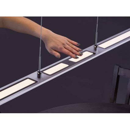 Dlhá strieborná lampa s tromi podsvietenými obdĺžnikovými plochami visí zo stropu. Ruka sa dotýka strednej plochy, ktorá slúži ako dotykový ovládač. Lampa je umiestnená nad tmavým povrchom.