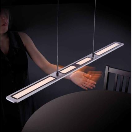 Dlhá LED lampa visí nad stolom. Žena pri stole pohybuje rukou pod lampou. Stôl a stolička sú v tmavom prostredí.