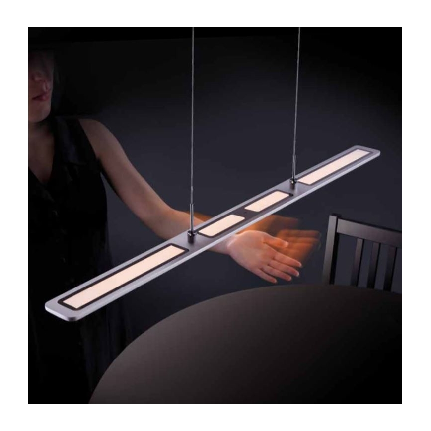 Dlhá LED lampa visí nad stolom. Žena pri stole pohybuje rukou pod lampou. Stôl a stolička sú v tmavom prostredí.