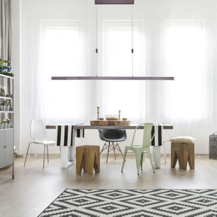 Dlhá lineárna lampa visí nad jedálenským stolom v svetlej miestnosti. Stoličky a stolík s ovocím sú umiestnené okolo stola. Podlaha je pokrytá čierno-bielym kobercom.