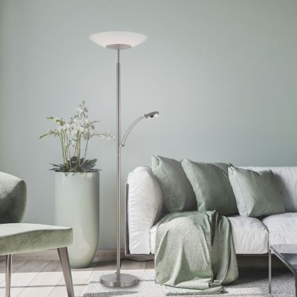 Stojacia lampa s nastaviteľnou čítacím svetlom stojí v obývacej izbe pri pohovke s vankúšmi a zelenou dekou. Vedľa je kvetináč s orchideami.