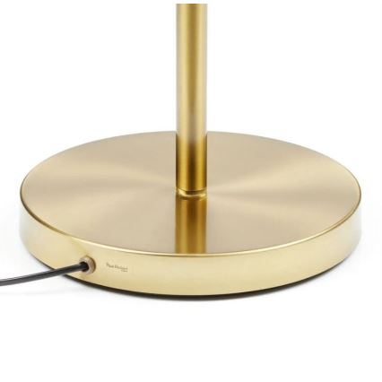 Zlatá kovová stojaca lampa s okrúhlou základňou. Na spodku základne je viditeľný nápis Paul Denleed. Stojí na svetlej ploche.