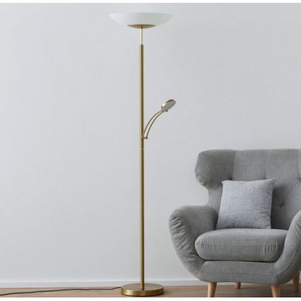 Zlatá stojacia lampa s dvoma svetlami stojí v miestnosti popri sivej kreslovej stoličke. Hlavné svetlo má široký biely tienidlo, menšie je otočné.