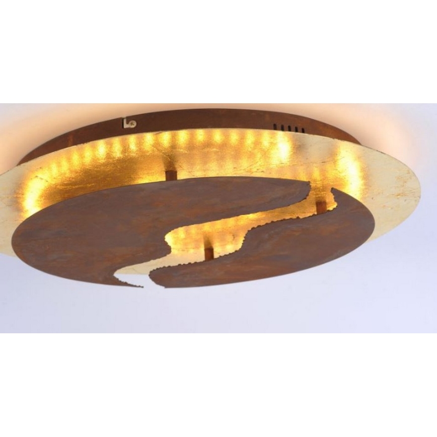 Okrúhle stropné svietidlo z hrdzavejúcej ocele a pozláteného kovu svieti. Svietidlo má vyrezaný abstraktný tvar. Je umiestnené na strope.