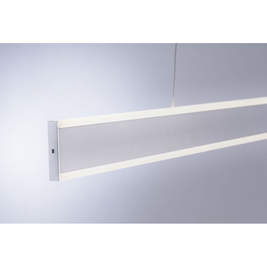 Dlhá obdĺžniková LED lampa visí zo stropu. Je strieborno-sivej farby s tenkým bielym LED osvetlením pozdĺž hornej a dolnej hrany. Svetlo je umiestnené v jednoduchom, neutrálnom prostredí.