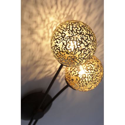 Dvojramenná nástenná lampa svieti. Dve guľové tienidlá z kovu s perforovaným vzorom vyžarujú svetlo. Lampa je pripevnená na stenu.