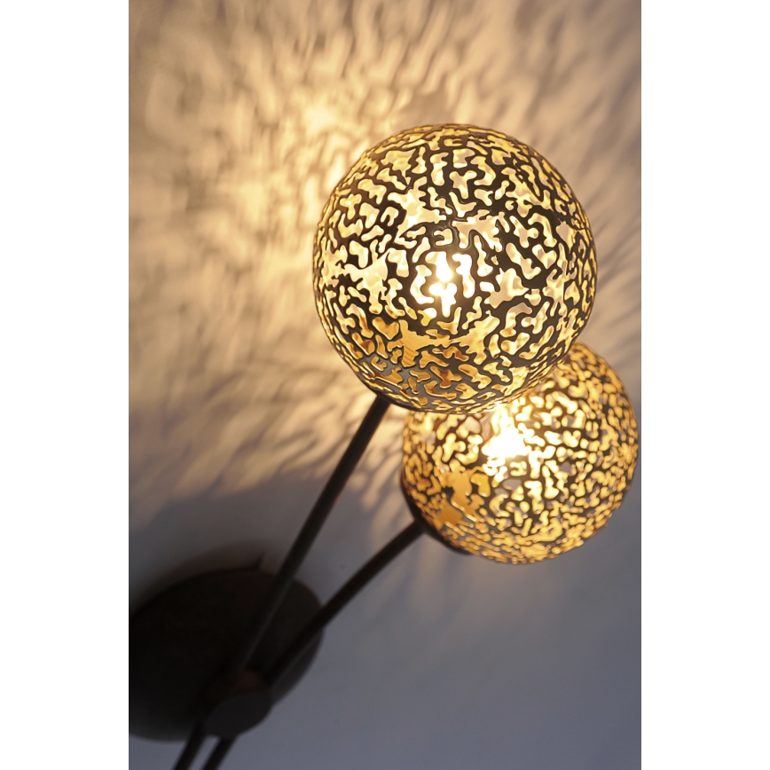 Dvojramenná nástenná lampa svieti. Dve guľové tienidlá z kovu s perforovaným vzorom vyžarujú svetlo. Lampa je pripevnená na stenu.