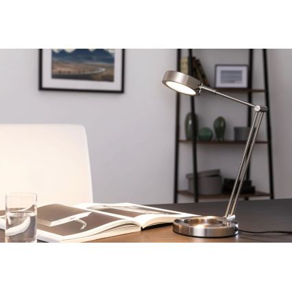 Stolová lampa s kovovou konštrukciou svieti na otvorenú knihu na drevenom stole. V pozadí je polica s dekoráciami a obraz na stene. V popredí je aj pohár s vodou.