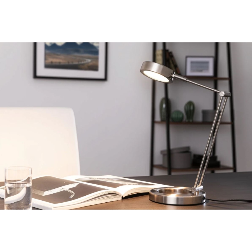 Stolová lampa s kovovou konštrukciou svieti na otvorenú knihu na drevenom stole. V pozadí je polica s dekoráciami a obraz na stene. V popredí je aj pohár s vodou.