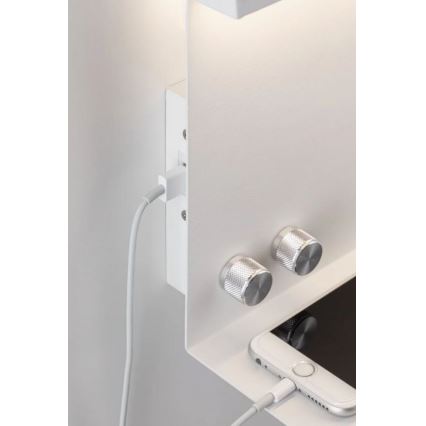 Biela nástenná polica s integrovanou zásuvkou a USB portom nabíja telefón. Na poličke sú dva strieborné otočné ovládače a svietiace svetlo nad ňou.
