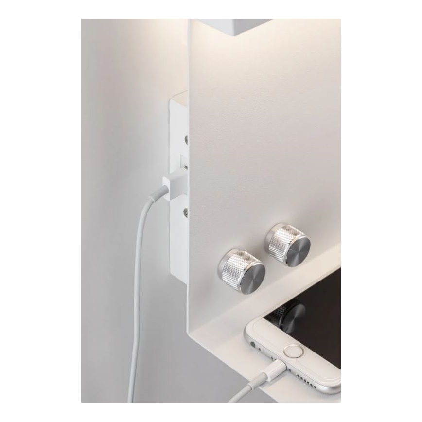 Biela nástenná polica s integrovanou zásuvkou a USB portom nabíja telefón. Na poličke sú dva strieborné otočné ovládače a svietiace svetlo nad ňou.