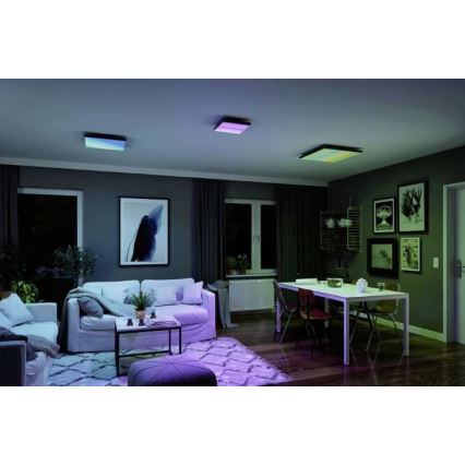 Štvorcové stropné LED panely svietia rôznymi farbami v obývacej izbe a jedálni. Svetlá sú umiestnené na strope. Prostredie tvorí zariadený byt s pohovkou, jedálenským stolom a nábytkom.