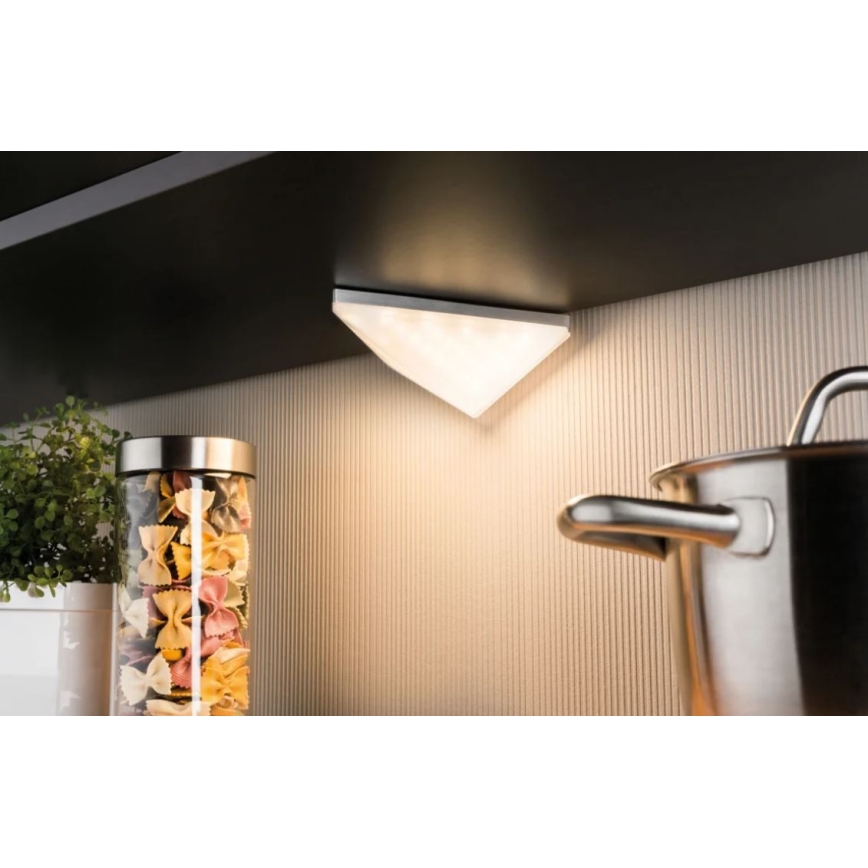 Trojuholníkové LED svetlo svieti pod kuchynskou linkou. Svetlo je pripevnené na tmavej poličke nad svetlohnedou stenou. V popredí je viditeľná nádoba s cestovinami a hrniec.