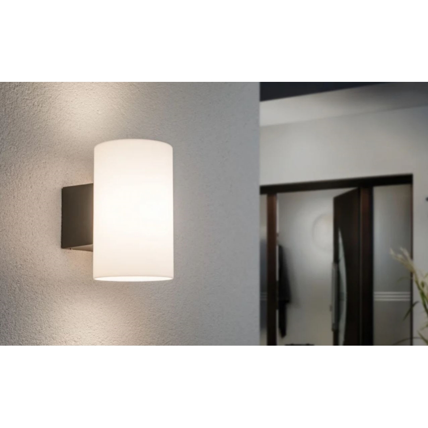 Nástenná lampa s valcovitým matným skleneným tienidlom svieti na svetlej stene. Lampa je tmavosivá. Pozadie ukazuje rozmazaný pohľad na chodbu s dverami.