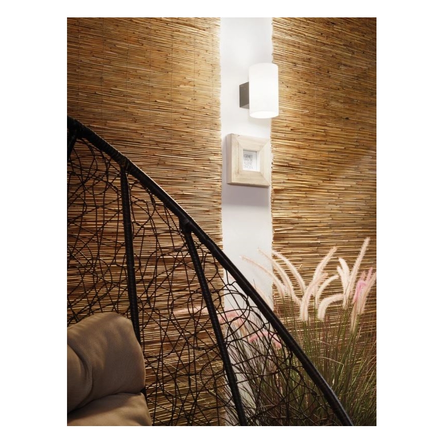 Nástenná lampa svieti na stenu s hnedým bambusovým obkladom. Vedľa nej visí malý rámik. V popredí je časť čierneho ratanového kresla.