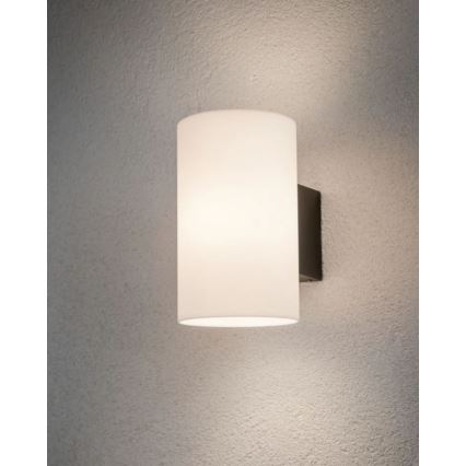 Nástenná lampa s valcovým matným skleneným tienidlom svieti na svetlej stene. Lampa má tmavosivý kovový základ.