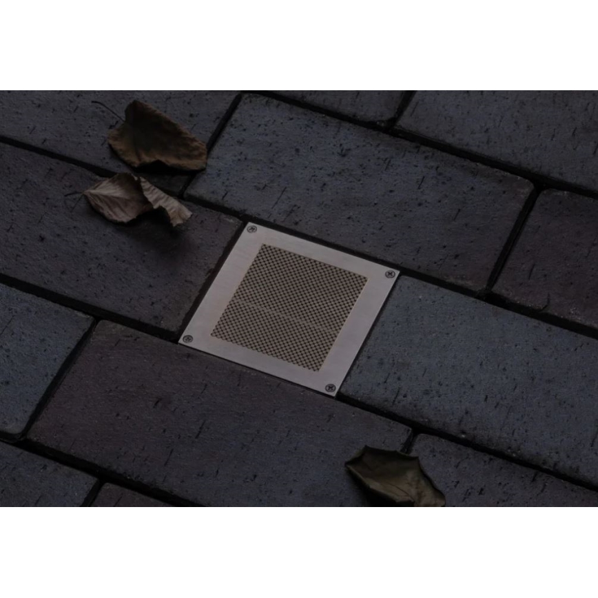 Štvorcový kovový ventilačný otvor je osadený do dlažby z tmavosivých tehál. V okolí ležia suché listy.