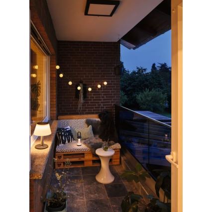 Stolová lampa svieti na balkóne zariadenom paletkovým nábytkom a vankúšmi. Okolitý priestor je osvetlený girlandou svetielok a stropným svietidlom. Balkón je v podvečer.