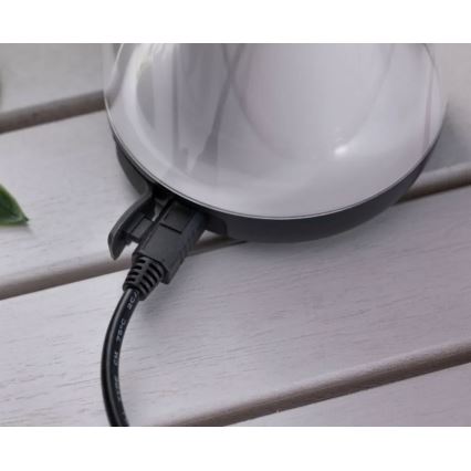 Okrúhle zariadenie s bielym vrchom a sivým okrajom je pripojené čiernym káblom k elektrickej sieti. Zariadenie leží na bielom drevenom stole.