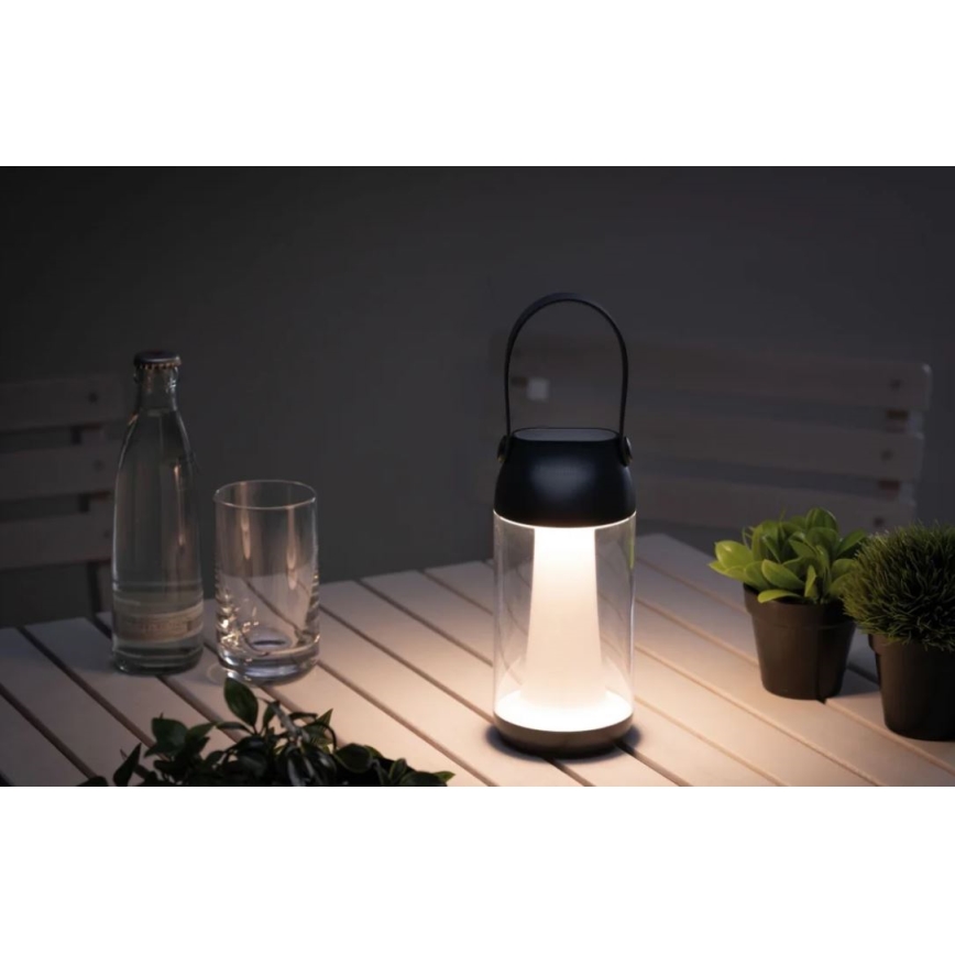 Prenositelná lampa svieti na bielom stole vonku. Lampa má čierny vrch s rúčkou a priehľadné sklenené telo. Okolitý priestor tvoria nápoje, rastliny v kvetináčoch.