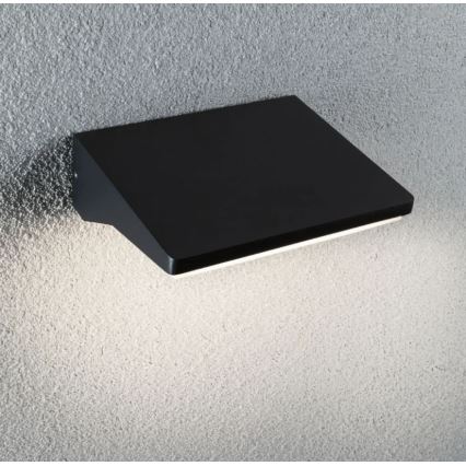 Čierna nástenná lampa svieti na štruktúrovanú stenu. Lampa má obdĺžnikový tvar so šikmým okrajom. Svetlo vyžaruje z jej spodného okraja.