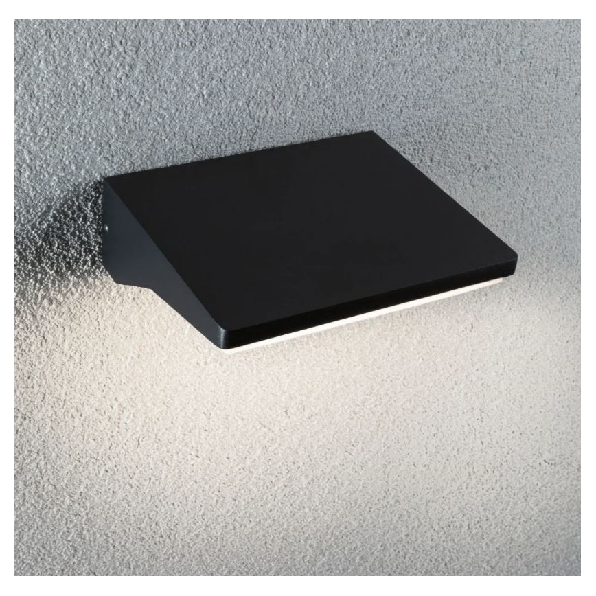 Čierna nástenná lampa svieti na štruktúrovanú stenu. Lampa má obdĺžnikový tvar so šikmým okrajom. Svetlo vyžaruje z jej spodného okraja.