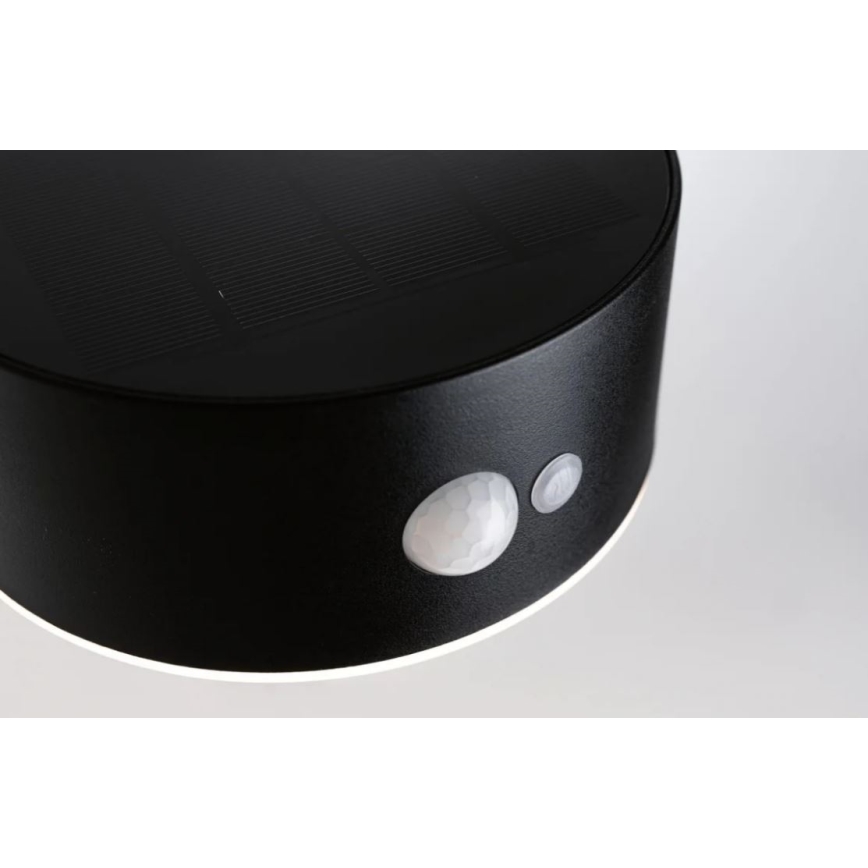 Solárny senzorový vonkajší svietidlo s detektorom pohybu je umiestnené na stene. Svietidlo je čierne, valcovitého tvaru, s horným solárnym panelom a na spodku je detektor pohybu a spínač.