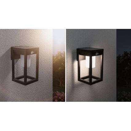 Solárna nástenná lampa s pohybovým senzorom svieti v tme. Je umiestnená na sivej stene v exteriéri. Lampa je čiernej farby, štvorcového tvaru s vnútorným mliečnym difúzorom.